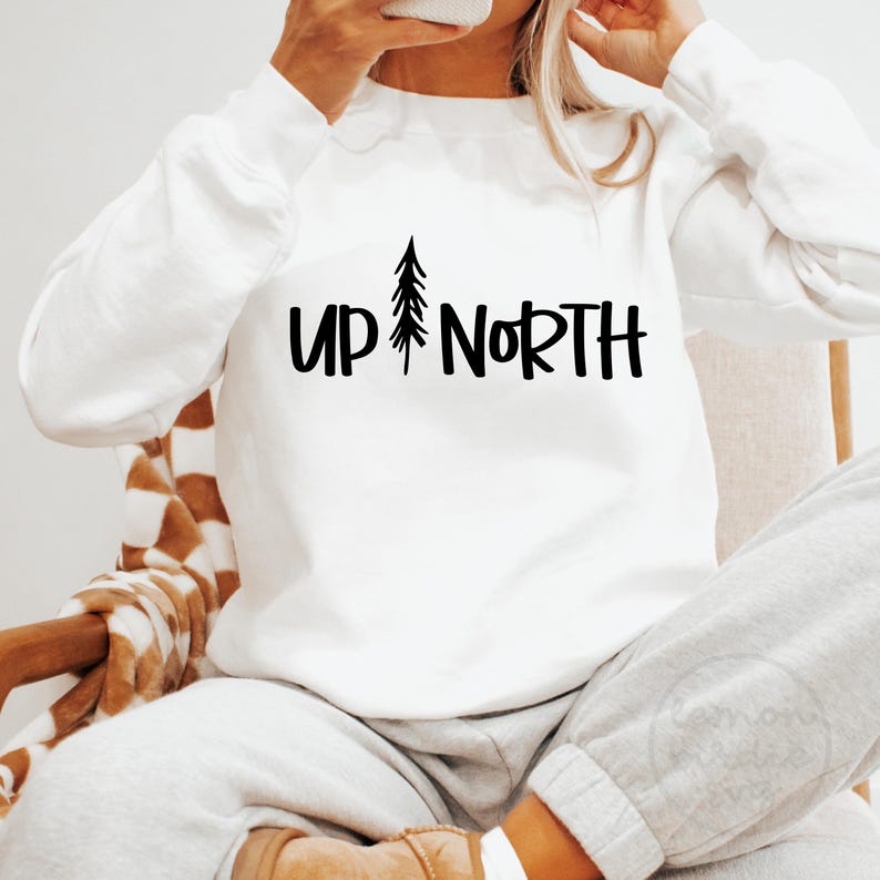 Op de afbeelding: Witte trui met zwarte tekst die "Up North" zegt en een kleine grafische afbeelding van een dennenboom.