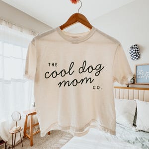 May include: A cream-coloured t-shirt with the text "THE cool dog mom co." in black script. The shirt is hanging on a wooden hanger. The background includes a bed, a small table, and a window with sheer curtains.