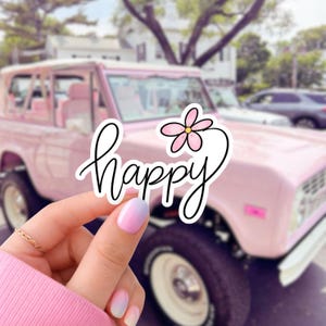 May include: A white sticker with the word "happy" in a cursive font, accompanied by a pink flower illustration. The sticker is held in front of a pink vintage car, with a blurred background of a street and buildings.