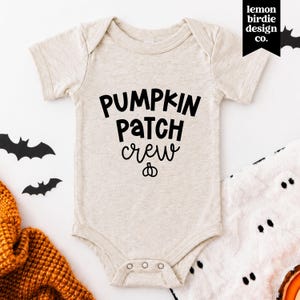 May include: A heathered beige short-sleeved baby bodysuit featuring the black text PUMPKIN PATCH CREW with a small pumpkin icon underneath. The garment has a round neckline and three snap closures at the bottom for easy access and secure fastening.
