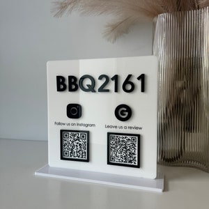 May include: A white acrylic sign with black lettering and graphics. The sign reads "BBQ2161" at the top, with Instagram and Google icons below, each accompanied by a QR code and text. The sign is on a white base.