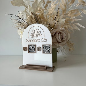 Op de afbeelding: Een witte standaard met een bruine basis, met een logo met de tekst "Sandpitt Co." en een gestileerde zonafbeelding. De standaard heeft twee QR-codes, een voor Instagram en een voor Facebook, met bijbehorende pictogrammen.