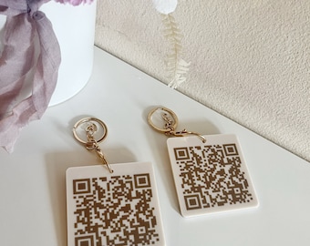 May include: Two white keychains with gold-colored QR codes printed on them. The keychains are on a white surface with a bouquet of pink and white flowers in the background. The text "LUXE DECOR BY LISA" is printed on the surface.