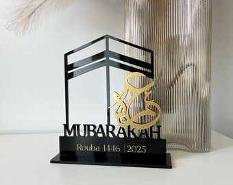 Peut inclure: Support en acrylique noir avec une découpe dorée de la Kaaba et le mot arabe "Mubarakah". Le support comprend également le texte "Rouba 1446 | 2025".