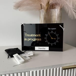 May include: A black acrylic sign with gold lettering reading "Treatment in progress" and a clock face that says "Re-open". Includes small white labels for days of the week. A black bag and decorative vase are also present.