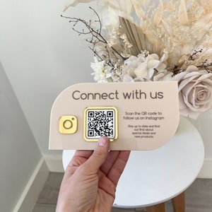 May include: A beige sign with the text "Connect with us" and a QR code. The sign also includes the text "Scan the QR code to follow us on Instagram. Stay up to date and find out first about special deals and new products."