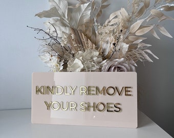 Peut inclure: Une pancarte rose avec des lettres dorées qui indique "KINDLY REMOVE YOUR SHOES". La pancarte est posée sur une surface blanche devant un bouquet de fleurs séchées.