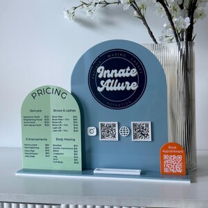 May include: A display board advertising esthetic services, featuring a blue arched panel with the Innate Allure logo and a green panel listing treatments. The sign includes a pricing menu for skincare, brows, lashes, enhancements, and body waxing. A vase of white flowers is in the background.