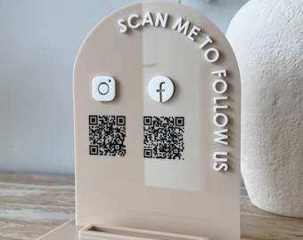 May include: An arched beige acrylic sign featuring the white text SCAN ME TO FOLLOW US, along with white Instagram and Facebook icons and two black QR codes. The sign is mounted on a rectangular beige base with a small raised rectangular frame.