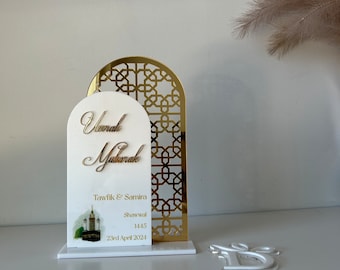 Peut inclure: Un support décoratif blanc et or avec un motif découpé au laser. Le support présente une plaque blanche avec le texte "Tawfik & Samira, Showwal 1445, 23rd April 2024" et le texte arabe "Umrah Mubarak".