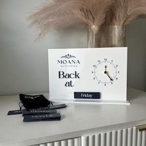 May include: A white desk clock with the text "MOANA ACTIVITIES Back at" and a clock face. Includes a black bag and several small black rectangular labels with the days of the week. A vase with decorative plumes is in the background.
