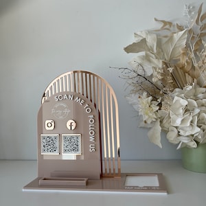 May include: A contemporary acrylic display stand in a neutral hue, featuring a rose gold arch design. The stand displays the text "SCAN ME TO FOLLOW US" and includes Instagram and Facebook icons with QR codes. A small rectangular tray and a "TAP HERE" section are also visible.