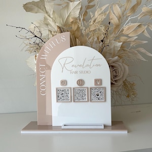 May include: A white and pale pink acrylic sign for a hair studio, with the text "Revelation Hair Studio" and social media icons with QR codes. The sign is decorated with dried flowers and the words "Connect With Us".