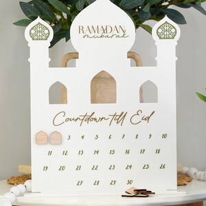May include: White wooden Ramadan countdown calendar shaped like a mosque with gold accents. The calendar has a crescent moon on top and the words "Ramadan Mubarak" written in gold. The calendar has numbers 1 through 30 for counting down to Eid.
