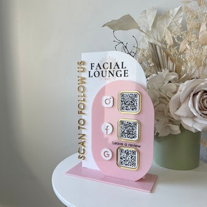 May include: A pink and white sign with the text "Facial Lounge OKC" and the words "Scan to Follow Us" written vertically on the side. The sign has three QR codes with icons for Instagram, Facebook, and Google. The text "Leave a review" is written below the Google QR code.