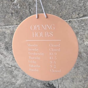 May include: A peach-coloured round sign with white text that reads "OPENING HOURS" and lists the days of the week and corresponding hours of operation. The sign is hanging from a string.