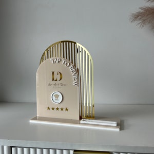May include: A gold and white stand with a sign that says "Tap to Review" and "L's Lips And Dripz by Brianna." The stand has a white NFC symbol and five white stars.