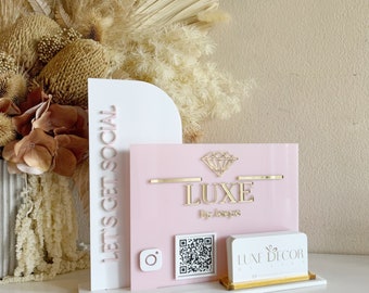 May include: A white and pink acrylic sign with the text "Let's Get Social" and "LUXE by Lisa". A diamond shape is above the text "LUXE". A white business card with the text "LUXE DECOR BY LISA" is in front of the sign.