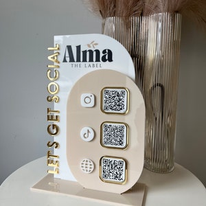 Peut inclure: Un présentoir en acrylique blanc et beige avec le texte "Let's Get Social" et "Alma The Label" en lettres dorées. Le présentoir comporte trois découpes circulaires avec des icônes de médias sociaux et des codes QR.