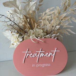 May include: A pink oval sign with white text that reads "treatment in progress". The sign is hanging from a white string and is surrounded by dried white flowers.