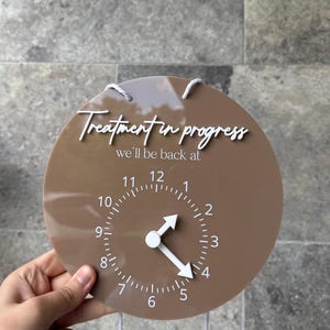 May include: A brown circular sign with the text "Treatment in progress, we'll be back at" in white script. The sign has a clock face with white hands and numbers. White string is attached at the top.