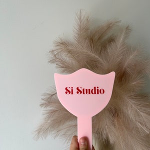 Op de afbeelding: Een roze handspiegel met de tekst "Si Studio" in het rood. De spiegel heeft een unieke vorm en wordt vastgehouden voor een achtergrond van gedroogde, veervormige planten.