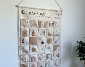 May include: A beige Ramadan Mubarak advent calendar hanging on a white wall. The calendar has 30 numbered pockets with illustrations of moons, lanterns, and mosques. The text "Ramadan Mubarak" is printed at the top, with "Let's Countdown to Eid" at the bottom.