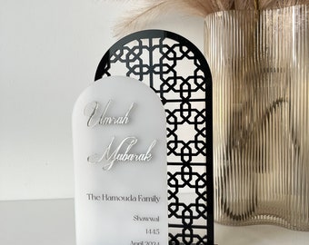 Peut inclure: Une plaque en acrylique blanche avec une calligraphie noire qui dit "Umrah Mubarak" et le texte "The Hamouda Family, Shawwal 1445, April 2024". La plaque est montée sur une base en acrylique noire avec un motif géométrique découpé au laser.