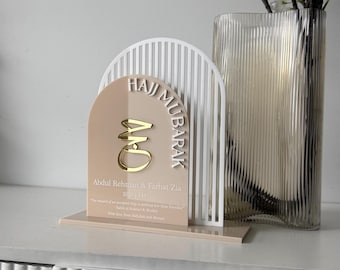 May include: An arched acrylic sign featuring a white slatted frame and a beige panel. The sign displays the gold text Hajj Mubarak, the names Abdul Rehman & Farhat Zia, the years 2026 | 1447, and a quote in white lettering on a rectangular base.