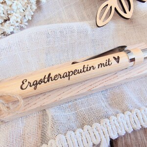 May include: A light-colored wooden ballpoint pen with a silver-toned metal clip and tip. The barrel is engraved with the German text "Ergotherapeutin mit" followed by a small heart symbol. The pen rests on a matching wooden rectangular base.
