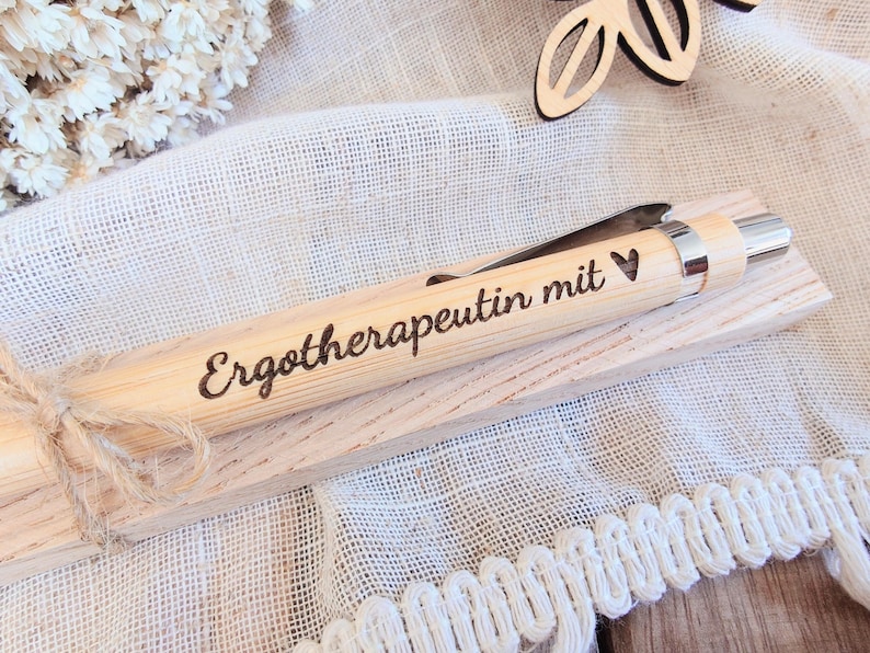 May include: A light-colored wooden ballpoint pen with a silver-toned metal clip and tip. The barrel is engraved with the German text "Ergotherapeutin mit" followed by a small heart symbol. The pen rests on a matching wooden rectangular base.