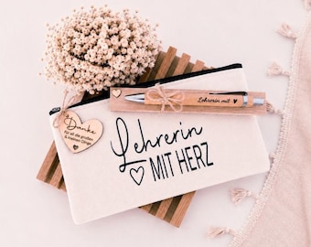 May include: A cream-colored canvas pencil case with the black text "Lehrerin MIT HERZ" and a heart icon. It includes a wooden pen engraved with "Lehrerin mit" and a heart, a heart-shaped wooden tag reading "Danke für all die großen & kleinen Dinge", and a bouquet of dried flowers.