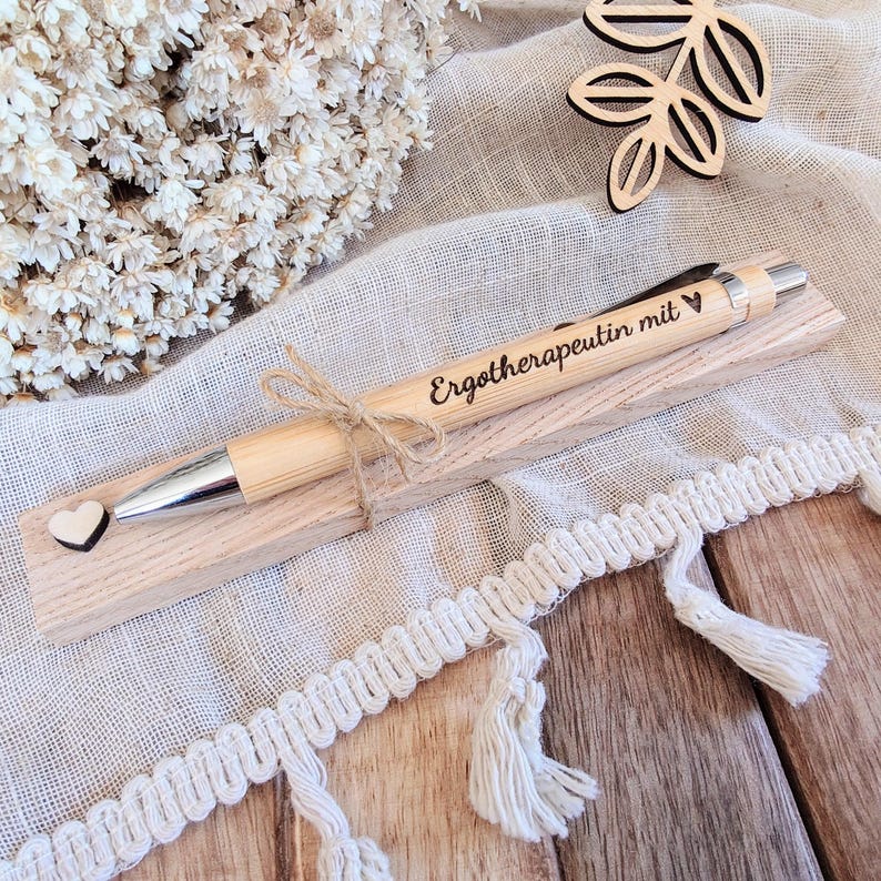 May include: A light-colored wooden ballpoint pen with a silver tip and clip, engraved with the text "Ergotherapeutin mit" and a heart symbol. The pen is tied with twine to a rectangular wooden base featuring a small wooden heart accent.
