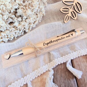 May include: A light-colored wooden ballpoint pen featuring a silver metal tip and clip, engraved with the text "Erotherapeutin mit" followed by a small heart symbol. A tiny wooden heart is attached to the pen, which rests on a wooden base.