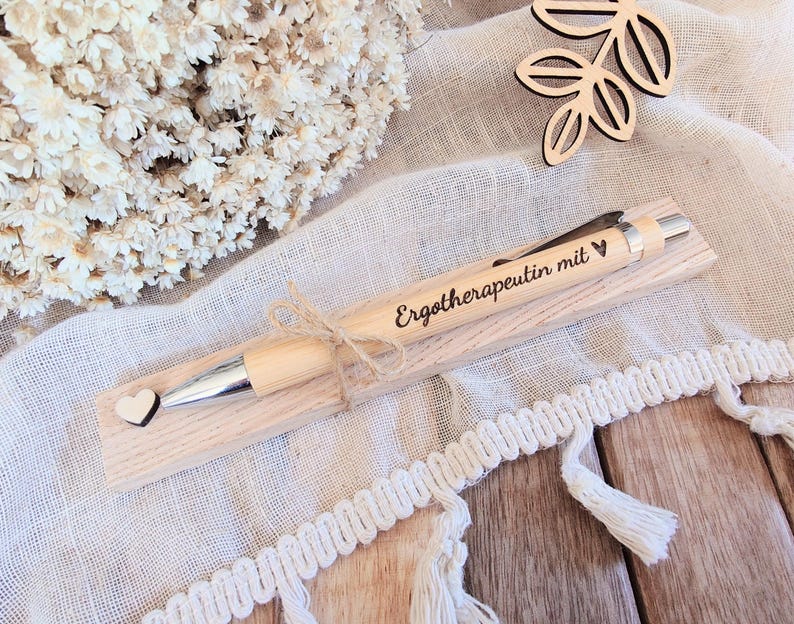 May include: A light-colored wooden ballpoint pen featuring a silver metal tip and clip, engraved with the text "Erotherapeutin mit" followed by a small heart symbol. A tiny wooden heart is attached to the pen, which rests on a wooden base.