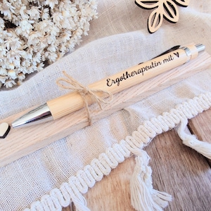 May include: A light-colored wooden ballpoint pen with a silver tip and clip, featuring the engraved text "Erotherapeutin mit" and a small heart symbol. The pen is tied with a thin jute twine bow and rests on a rectangular wooden base with a small heart.