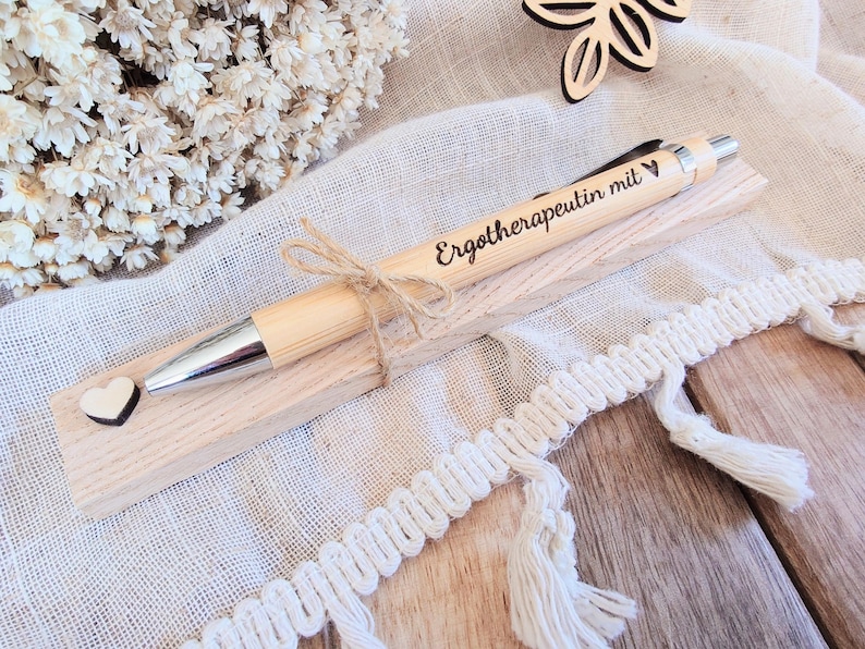 May include: A light-colored wooden ballpoint pen with a silver tip and clip, featuring the engraved text "Erotherapeutin mit" and a small heart symbol. The pen is tied with a thin jute twine bow and rests on a rectangular wooden base with a small heart.