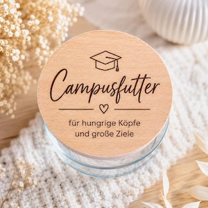 Könnte beinhalten: Ein runder, heller Holzdeckel mit einer lasergravierten Doktorhut-Ikone, einem kleinen Herz und dem Text Campusfutter für hungrige Köpfe und große Ziele. Der Deckel liegt auf einem klaren Glasbehälter mit einem glatten, kreisförmigen Rand.