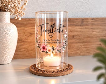 Puede incluir: Un portavelas cilíndrico de cristal transparente descansa sobre un posavasos de corcho redondo. El cristal presenta un diseño de corona floral, un pequeño corazón y el texto negro Trostlicht, Für stille Gedanken und liebe Erinnerungen. Una vela de té encendida está dentro.