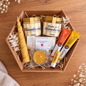 Könnte beinhalten: Eine sechseckige Geschenkbox mit zwei 500g Honiggläsern mit der Aufschrift Blutenhonig und Wald-und Blutenhonig, einer hohen spiralförmigen Bienenwachskerze, einer kleinen runden Kerze, einem Stück Seife und zwei Tuben Lindesa Hautschutzcreme.