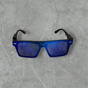May include: Blue sunglasses with a black frame and a bull logo. The lenses have a reflective blue tint. The sunglasses have a modern, rectangular shape.