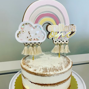 May include: A white cake with a rainbow, cloud, and tassel topper. The topper says "emiliana one".