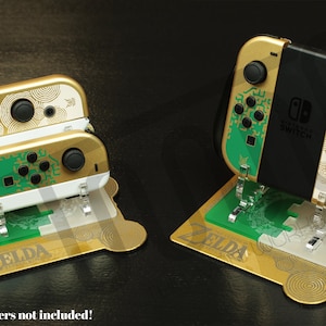 Pode incluir: Controladores Nintendo Switch Joy-Con dourados e verdes com um design temático de Zelda. Os controladores estão em um suporte dourado com um logotipo verde de Zelda. "Controladores não incluídos!" está escrito em vermelho na parte inferior da imagem.