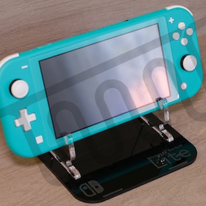 Puede incluir: Una consola Nintendo Switch Lite turquesa se muestra en un soporte negro. El soporte tiene las palabras "Nintendo Switch Lite" impresas en él.