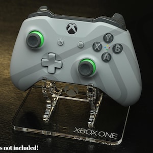 May include: A light gray Xbox One controller with green accents, resting on a clear acrylic stand. The controller features the Xbox logo and the letters A, B, X, and Y. The stand is labeled "XBOX ONE".