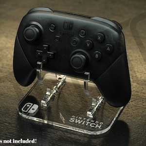 Puede incluir: Un mando Nintendo Switch Pro negro sobre un soporte de acrílico transparente. El soporte tiene el logotipo y el texto de Nintendo Switch. El mando tiene botones etiquetados X, Y, A y B. El soporte está sobre una superficie oscura.