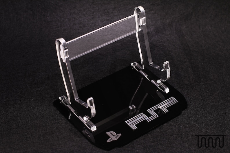 Op de afbeelding: Een transparante acrylstandaard met een zwarte basis. De basis heeft het PlayStation-logo en de tekst "PS2" erop gedrukt. De standaard is ontworpen om een ​​videogamesysteem te houden.