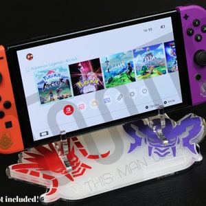 Könnte beinhalten: Ein transparenter Acryl-Ständer mit einem roten und violetten Design, das einen Videospielcharakter zeigt. Der Ständer hält eine Nintendo Switch-Konsole mit einem violetten und orangefarbenen Controller auf jeder Seite. Der Bildschirm der Konsole zeigt das Spiel "Pokémon Legends: Arceus". Der Text "THIS MAN" ist auf dem Ständer aufgedruckt.