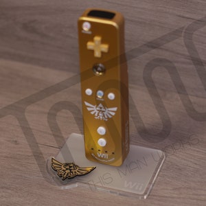 Puede incluir: Un mando de Wii dorado con un símbolo Triforce blanco y las palabras "Wii Remote" impresas en él. El mando está sentado en un soporte de plástico transparente con las palabras "This Man" impresas en él.