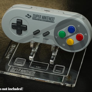 May include: A gray Super Nintendo controller rests on a clear acrylic stand. The controller features a directional pad, buttons in blue, green, red, and yellow, and the Super Nintendo logo. The stand has the Super Nintendo logo and button labels.